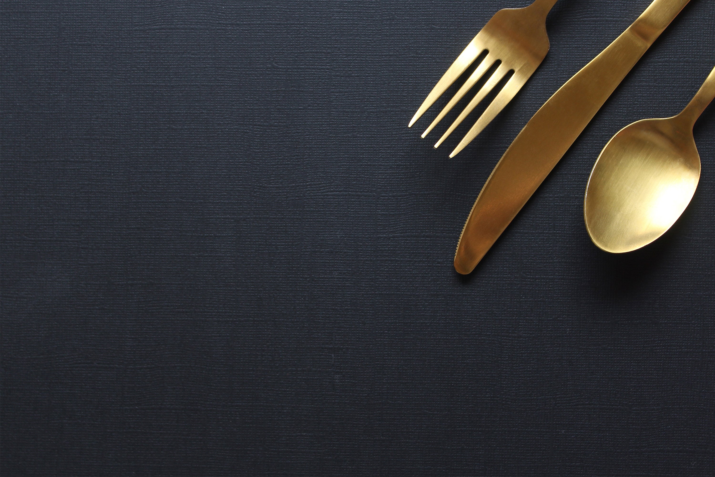 Een elegante goudkleurige bestekset, bestaande uit een vork, mes en lepel, netjes gerangschikt op een donkerblauwe, textielachtige achtergrond.   2 / 2      Was this response better or worse?  Better  Worse  Same